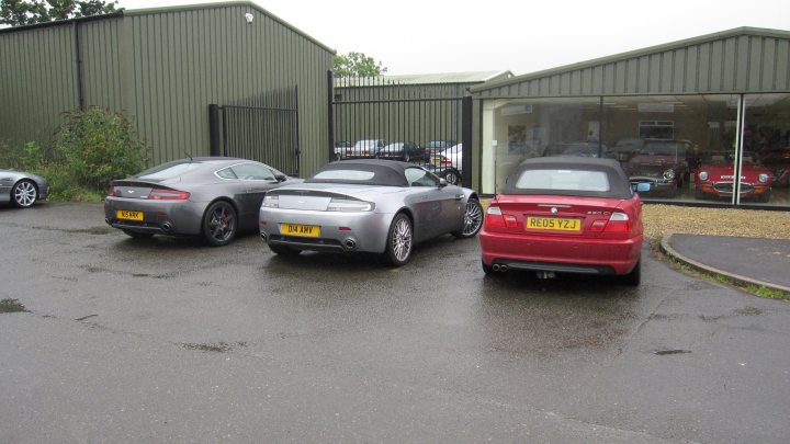 Pistonheads - The image depicts a parking lot with several cars parked beside each other, in front of a building or warehouse. The lot is wet, indicating that it has recently rained. The cars vary in color and model but are aligned parallel to a fence or wall, indicating an organized parking arrangement. In the background, there is a building with a reflective surface, possibly a window, that is visible through the wet pavement. A grassy area can also be seen behind the cars.