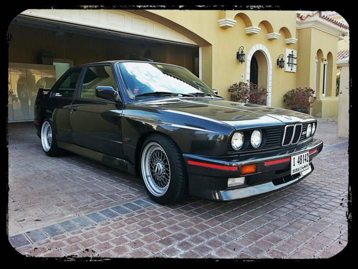 Pistonheads - The image features a sleek black BMW parked in a driveway. The car is positioned in front of a house with a beige exterior. The license plate on the car reads "T92B". The car appears to be in a healthy condition, and its position suggests it is in a residential area. The driveway itself has a tan hue, and there are potted plants on either side, adding a touch of greenery to the scene. The car features chrome trim and alloy wheels, adding to its luxurious appearance.