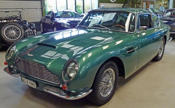 Pictures from Aston Workshop, Beamish - Page 1 - Aston Martin - PistonHeads - The image displays a vintage Aston Martin car parked indoors, possibly in a garage or showroom. The car is a deep, rich shade of green and appears to be in very good condition, suggesting it may be a classic model with well-maintained defenses. The vehicle's round headlights, classic grille, and round spoke wheels are characteristic of the Aston Martin design ethos of its time. The red reflector in the taillight is still visible, indicating either that the car is new or that it has been meticulously preserved. The setting is illuminated by bright overhead lights, highlighting the classic car's striking color and polished condition.