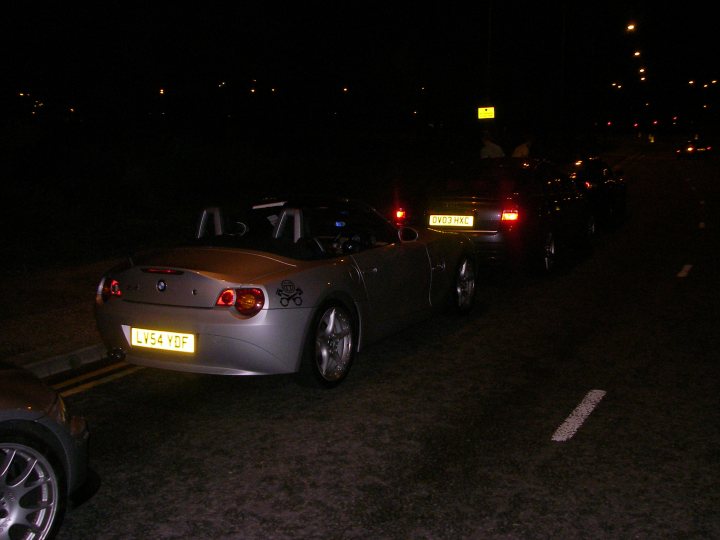 My Z4, may she surely be missed... - Page 1 - Readers' Cars - PistonHeads - The image depicts a dimly lit street at night, with a row of cars parked on the right side of the road. The lead car is a BMW, distinguishable by its emblem and license plate. The BMW is next to a car with its brake lights on, and the car in the front is casting light onto the road with its headlights dimly lit. The scene conveys a tranquil evening in a city or town environment.