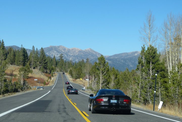 Pistonheads - The image showcases a winding stretch of a highway that is positioned between two ranges of mountain peaks. The sky above is a clear blue, suggesting a calm and possibly sunny day. Cars can be seen traveling on the road in different directions, indicating active traffic. Trees and bushes line either side of the road, contributing to a scenic view that combines natural beauty with a sense of motion. The landscape is devoid of any noticeable urban elements, enhancing the feeling of a rural route.