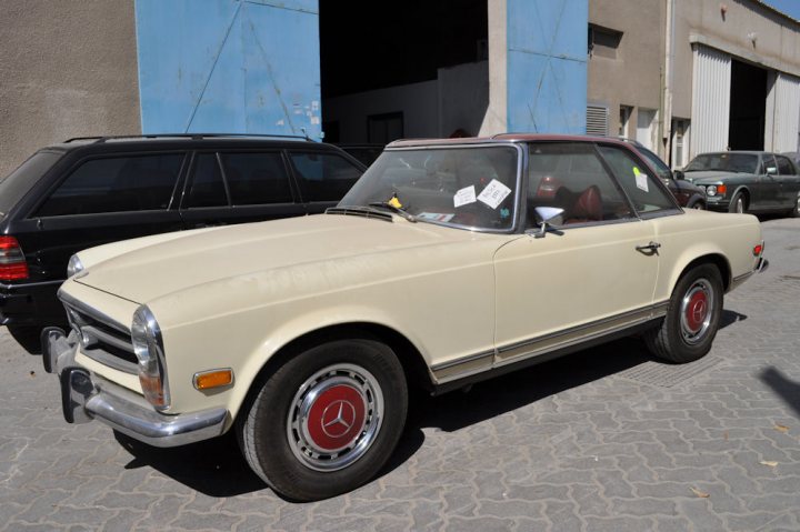 Pistonheads - The image displays a white beetle car parked on the street during the day. The car is positioned in front of a package shop, as indicated by the white door. The side view of the car shows a classic design with matching red hubcaps. The street appears to be a parking lot with some commercial buildings visible in the background.