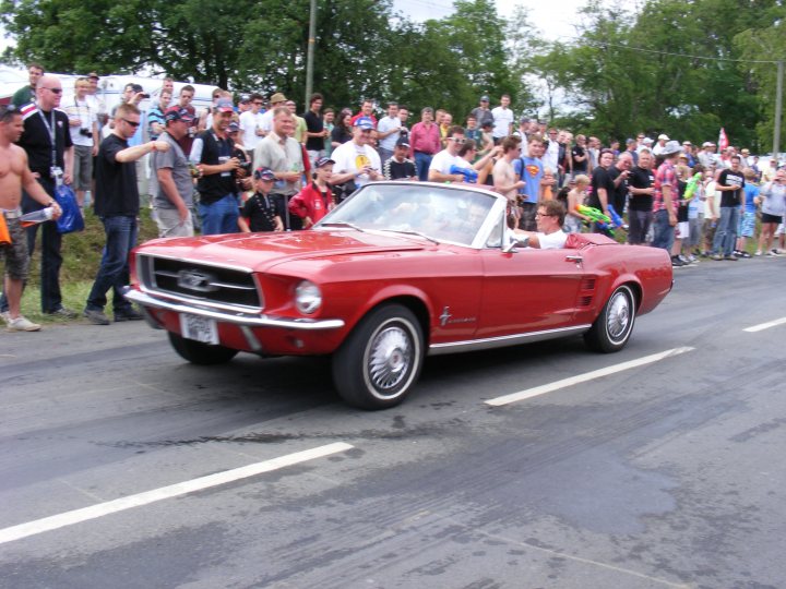 Pistonheads Classics - The image captures a lively street scene. At the center of the action is a red Mustang convertible, possibly a rally classic, driving down the street with a convertible top down. A large crowd of people are lined up along the sides of the street, watching the Mustang with keen interest. The crowd consists of a mix of individuals, some flying flags and others waving, adding to the festive atmosphere. The Mustang is parked on a wet road, suggesting it may have just raced or is part of an auto show. The presence of a soccer ball chimney suggests that there might be inflatable bouncy houses nearby, hinting at a community event or parade.