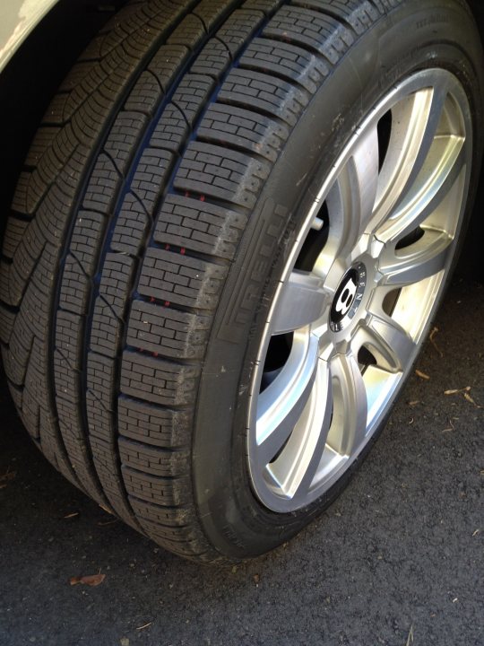 I’ve put my football boots on & bought a Bentley GT Speed - Page 1 - Rolls Royce & Bentley - PistonHeads - The image shows a close-up view of a car tire. The tire is positioned at an angle, revealing its tread pattern and the tire's sidewall. It appears to be a standard car tire with a silver hubcap. The ground beneath the tire is paved, and there are small, dry leaves scattered around near the rim.