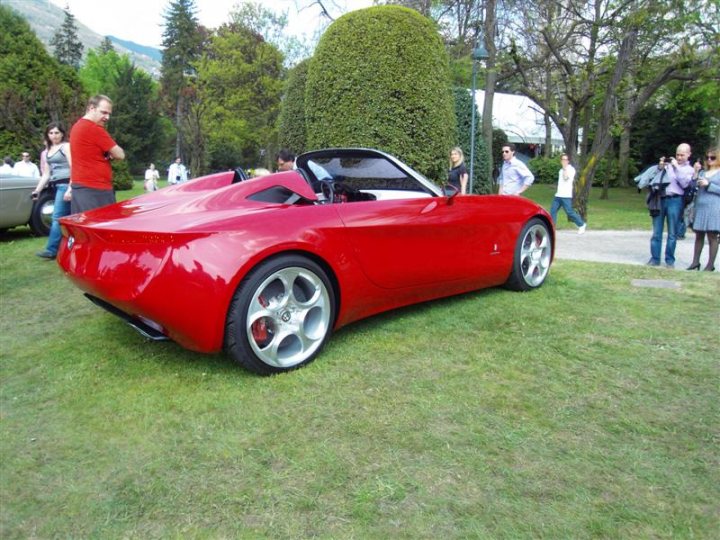 Deste Pistonheads Villa Concoursporn - The image features a bright red sports car parked on a grassy area, drawing attention from onlookers. The car is a convertible with the top down, allowing a clear view of its sleek design. It's a sunny day as indicated by the shadows of those standing around the car, who are observing and admiring the vehicle. The setting appears to be a park or a public space where people can gather and enjoy the outdoors.