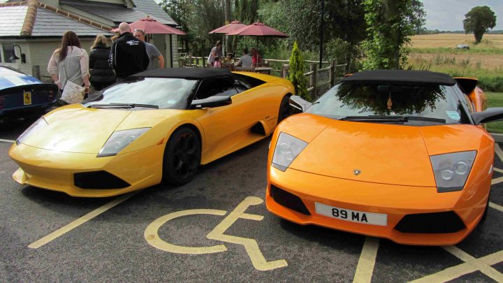 Hare Breakfast Meet - August - Page 1 - Kent & Essex - PistonHeads - The image shows two fast, shiny sports cars parked side by side on a street. The car on the left is a yellow-toned vehicle, while the one on the right appears to be a vibrant orange color. Both cars have sleek designs with aggressive front ends and prominent grilles, indicative of their high-performance status. There are people present on the sidewalk, suggesting a bustling urban environment. The street is dotted with parked cars further back, and there are some umbrellas visible, possibly attached to outdoor seating areas of local businesses.