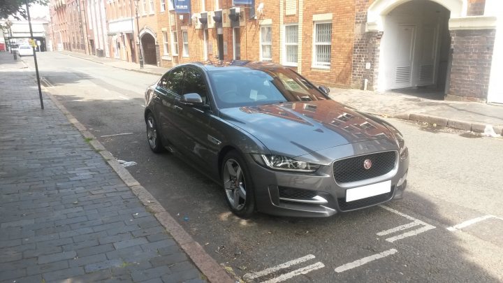 Jaguar XE R-Sport [180] Ammonite Grey with Jet/Oyster Seats - Page 1 - Readers' Cars - PistonHeads - The image captures a vibrant city scene with a black Jaguar car parked on the street. The car is sleek, with the Jaguar emblem prominently visible on the front grill. It's parked neatly next to a yellow line, indicative of a parked car area. The street is lined with brick buildings that give off a historic charm. The area appears well-maintained, with a clean and tidy aesthetic. The car seems to be the central focus of the image, drawing attention to its shiny surface and prominent logo, emphasizing its elegance and luxury.