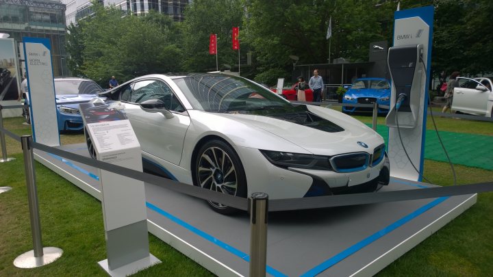 Tesla and i8 at Canary Wharf motorshow this week - Page 1 - EV and Alternative Fuels - PistonHeads - The image showcases an impressive display of a white BMW car, which is a two-door model renowned for its sleek design and performance. The car is surrounded by a protective blue barrier, indicating a formal event or show. Behind this barrier, a detailed information stand is available, likely providing detailed specifications and features of the car. Surrounding the display area, there are a number of visitors observing the car, contributing to the lively atmosphere of the event. The car's position on grass suggests that this may be an outdoor exhibit or show.