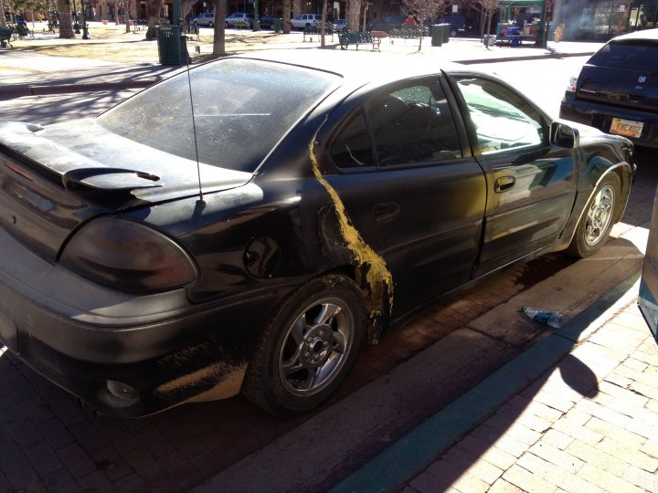 Worst bodge you have seen - Page 12 - General Gassing - PistonHeads - The image shows a black sedan parked on the side of a road. The most noticeable feature is a large, yellow substance splattered along the side, reaching towards the rear of the car. The collision appears significant, with parts of the car's bodywork damaged. The car's window glass has partially shattered and is scattered around the vehicle. The surrounding area seems to be a sidewalk next to a building and a street scene with clear skies and trees. The white of the car and the yellow of the substance create a stark contrast.