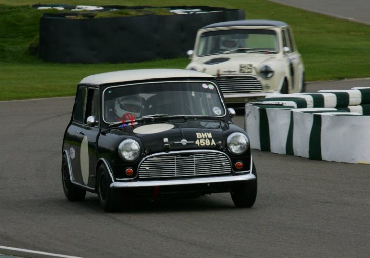 Masters race series, 10th March - Page 1 - Goodwood Events - PistonHeads - The image shows a black and white vintage car racing on a track. The vehicle is prominently placed in the foreground, with its headlights and grill clearly visible. There are two barrel-shaped objects near the car, possibly traffic barriers or markers on the track. Behind the car, there is a simple, unadorned barrier, and to its right, another vintage car is visible, appearing to be in motion. The setting suggests an old-fashioned racing event or a historical show.