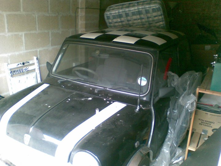 Restoration Body Mini Pistonheads Shop Needed - The image shows a black Mini Cooper car parked indoors, possibly a garage, with a brick wall in the background. The unique feature of the car is a checkered pattern on the hood, which is not standard for a Mini Cooper. The car appears to be a classic or vintage model, as indicated by its design. There's an open storage compartment in the back, and the car is covered with a clear plastic shelter, which is a common feature for vehicles stored indoors. It is daylight outside the garage.