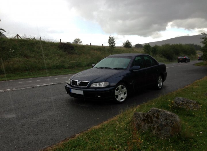 2002 Omega 3.2 V6 Elite Saloon - Page 1 - VX - PistonHeads - The image shows a dark blue compact car parked beside a road during what appears to be rainy weather, given the sky and the potential presence of water droplets on the car and the ground. In the background, there are trees lining the road, with a grassy area to the right and a fence to the left of the car. Another vehicle is visible in the distance. The scene suggests a quiet, rural setting.