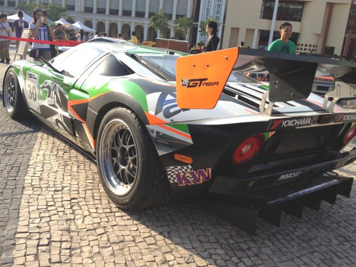 A car is parked on the side of the road - Pistonheads - The image features a dynamic racing scene, dominated by a sleek and vibrant racing car. The car is primarily black, with splashes of color from sponsors such as Striker, Mackerel, and Kwik, and the green and red colors of a checkered flag. The car is parked on a cobblestone street, set against the backdrop of a cityscape with buildings and people visible in the background. A man is seen walking behind the car, his figure slightly smaller due to the camera's perspective. The overall atmosphere suggests an event related to motor racing.