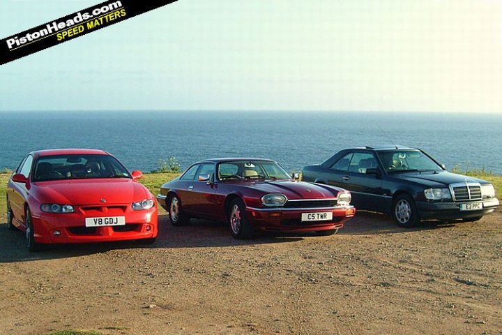 Side to Side: The Sunset to Sunrise Challenge (2011) - Page 1 - South West - PistonHeads - This image showcases a scenic setting with three cars parked on a dirt ground alongside a coastal road. The cars in the foreground are positioned in a triangle, with their fronts away from the viewer, facing the water. A red sports car and a black sedan can be seen; the third car, a deep red sports vehicle, is situated behind the other two. The backdrop is a vast body of water, which appears to be calm, reflecting a sense of tranquility. In the top left corner of the image, there is a black banner with white text that reads "PistonHeads.com SPEED MATTERS," indicating the source of the image and suggesting a theme of automotive speed or interest.