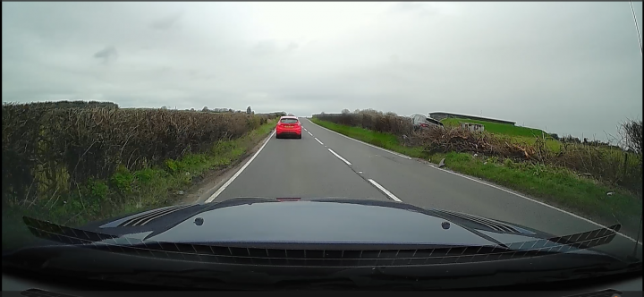 Pistonheads - The image shows a rear-view mirror mounted on the top left corner of the car. In the mirror, there's a reflection of a rural landscape with fields and a country road. A car is visible in the mirror as it drives away from the viewer's perspective. There's also a glimpse of a sky that appears to be overcast. The overall tone of the image is muted due to the grey weather conditions and the glass of the rear-view mirror.