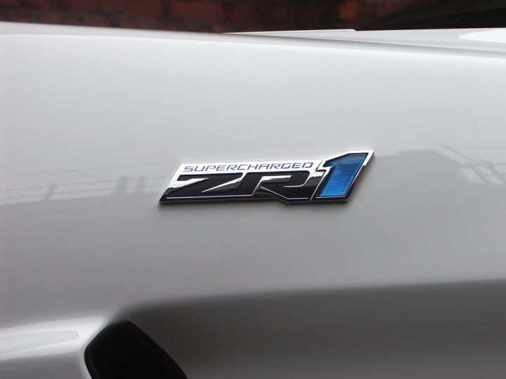 How about some ZR-1 photos to cheer you up? - Page 1 - Corvettes - PistonHeads - The image focuses on a gray patina of a vehicle, captured in close-up. On the top left corner, there is a distinctive blue and white logo of the ZR1, a supercharged car. The logo, standing out against the gray background, represents the car's unique identity. The car itself is parked, and other vehicles are captured in the background. The image suggests that the ZR1 is parked amidst other vehicles, perhaps at a show or gathering.