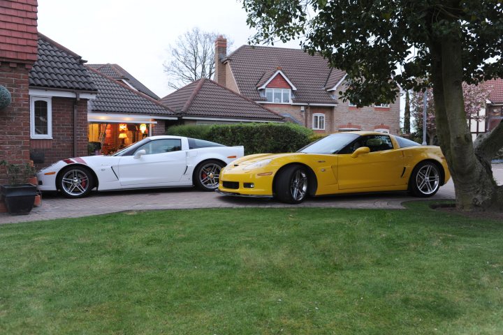 Clear side markers C6 - anyone got some? - Page 1 - Corvettes - PistonHeads - This image captures a tranquil outdoor scene featuring a house and driveway. The driveway is adorned with two luxury sports cars, one yellow and one white, both parked neatly. Directly behind these vehicles is a stately red brick house with a white fenced garage. To the side, there's a black trash can and a potted plant, adding to the suburban ambiance. The sky is blue, and the grass is a lush green.