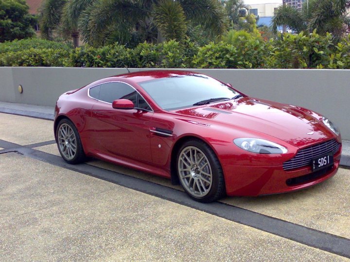 Phers Pistonheads Aussie - The image features a striking red Aston Martin car parked on the side of a road. The car is positioned at a slight angle to the camera, highlighting its sleek design and shiny finish that reflects the environment around it. It's in excellent condition, with well-maintained rims and a polished exterior. The background is relatively subdued, focusing attention on the car itself, but there are hints of greenery and a clear sky. The image does have a watermark, indicating it may be from a photographer's portfolio, but the specific details are not legible.