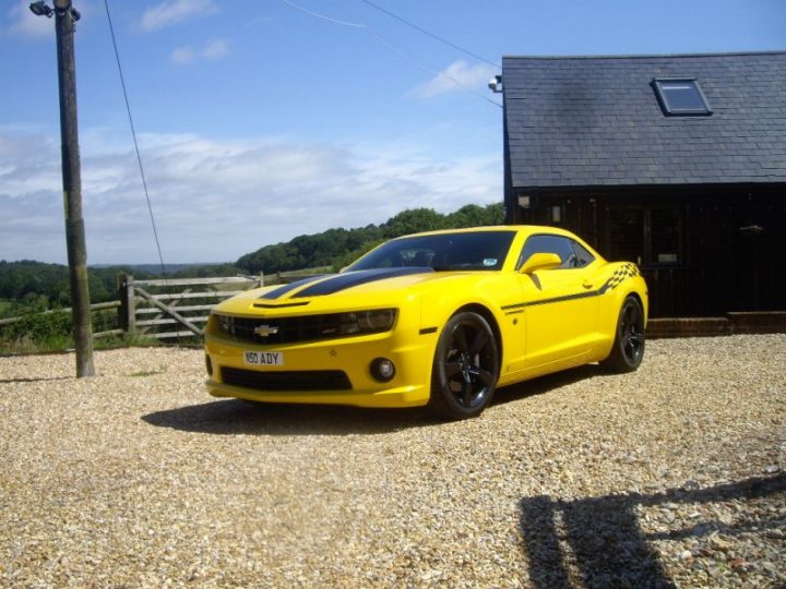 Any ZL1s in the UK? - Page 1 - Yank Motors - PistonHeads - This image features a vibrant yellow Chevrolet Camaro sports car parked on a pebbly surface in front of a dark-colored building with a gray roof. The car's striking yellow color, characteristic headlights, and sleek bodywork distinguish it as a Camaro. The sky above is blue, suggesting that the picture was taken on a clear day. The driveway leading to the building is lined with lush greenery, adding a touch of nature to the scene.