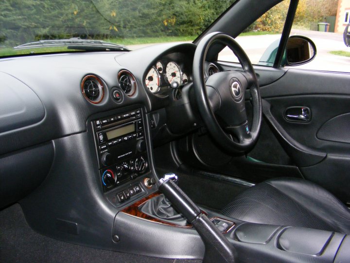 Show us your MX-5 .  56k run away - Page 36 - Mazda MX5/Eunos/Miata - PistonHeads - The image presents a sleek and well-maintained black car interior. Mounted on the center console is a stereo system with various buttons and dials, indicative of a high-end audio setup. Directly above this, a steering wheel with a silver logo, possibly representing the manufacturer, is visible. In the foreground, a comfortable-looking black car seat stretches across the frame of the image, providing a sense of luxury and refinement. The reflection of the steering wheel and some of the dashboard features can be seen in the side mirror, adding depth to the image.