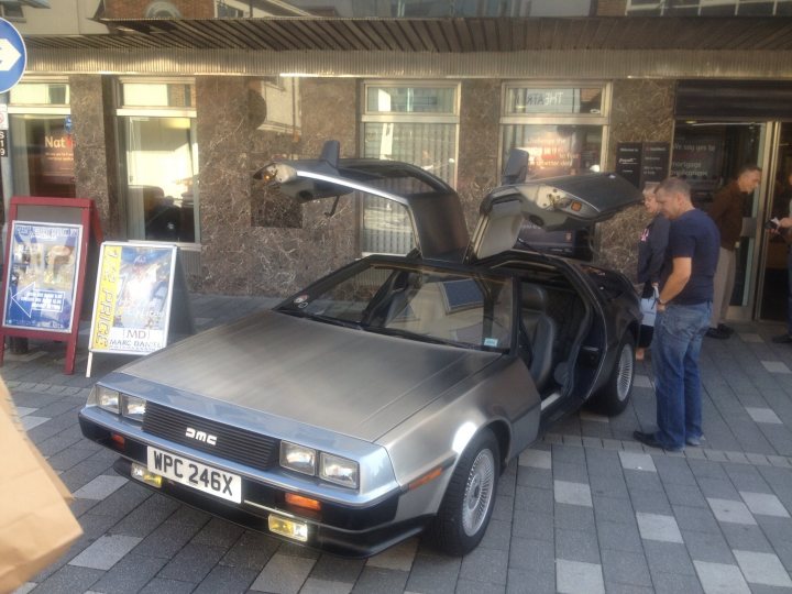 A car is parked in front of a building - Pistonheads - The image features a parked silver DeLorean, which is prominently displayed in a street-side driveway. The car is from the 1980s and is open, allowing two people to inspect its interior. The driveway is near a building with closed doors, and there's a blue sign with an arrow pointing downwards. The scene appears to be set in an urban area.