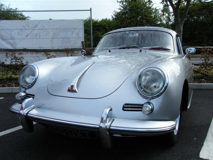 Breakfast Meeting, The Hare, Roxwell 4th June - Page 1 - Kent & Essex - PistonHeads - The image depicts a classic silver sedan parked in a parking lot. The car appears to be an older model, possibly a convertible, judging by the curve of the roofline and the large grille in the center of the car. The vehicle shines in the light, indicating it is in good condition. The parking space is neatly outlined with painted markings on the asphalt surface. In the background, you can see a construction site with a tall fence and a partially obscured tree. There are no people visible in the busy outdoor setting.