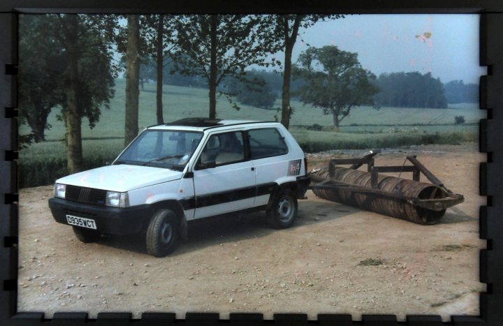 Fiat Panda 4x4 Mk 1 Fire.  A  Classic Car - Page 1 - Classic Cars and Yesterday's Heroes - PistonHeads - The image is a photograph depicting a small white truck situated on dirt, with a large cylindrical object next to it. The truck has a compact design, and there's a spare tire strapped to its rear, bolted onto a fabrication that appears to be homemade. Behind the truck, there's a peaceful rural setting with trees and grass in the background. In the top right corner, there's a faded or crying out graphic that may indicate some distress or urgency.