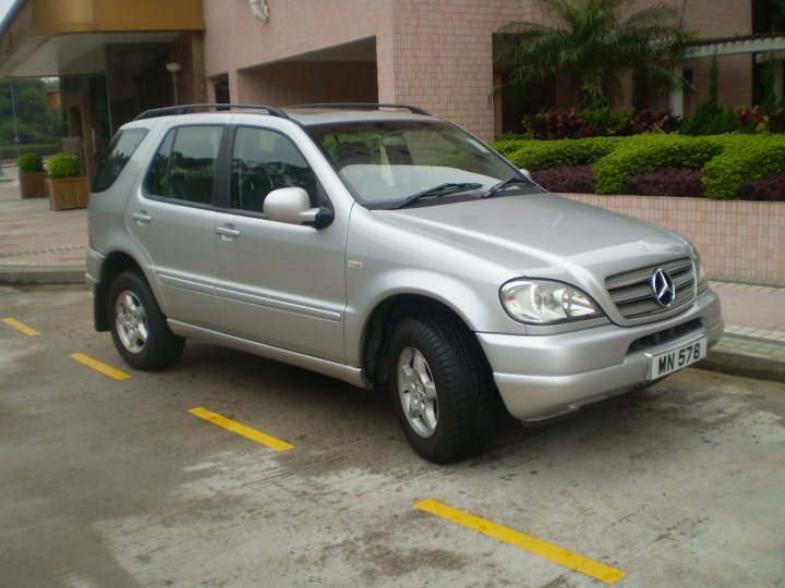 buying and driving a car in HK - Page 1 - Asia - PistonHeads - The image shows a silver Mercedes-Benz SUV parked in front of a large, multi-storied building. The car is parked diagonally across two yellow parking lines. The building has a pink facade with some green shrubbery visible around its entrance. No people are visible in the photo. The setting suggests a commercial or possibly a residential area with well-manicured urban landscaping. The car is clean and well-maintained.