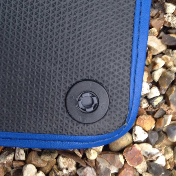 Floor Mats - Page 1 - Gallardo/Huracan - PistonHeads - The image provides a close-up view of a footbed mat with a black top and a blue trim. A circular button is attached to the footbed, which appears to be lined with a blue material that matches the trim of the mat. The background consists of rocks and pebbles, adding a naturalistic setting to the scene. The mat is laid flat on the rocky surface, suggesting it might be intended for use outdoors, perhaps as a seating area or for use during outdoor activities.