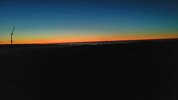 The "Photos From Today's Ride" thread... - Page 277 - Pedal Powered - PistonHeads - This image captures the breathtaking vista of a sunset. The sky is a canvas of colors, with hues of blue gracefully transitioning into vibrant orange and fiery red. The sun, partially hidden behind the horizon, casts a warm glow across the entire scene. Above the horizon, it's easier to see the silhouette of a flagpole and power lines, adding depth and contrast to the scene. The landscape at the bottom appears to be a mix of green and brown, suggesting a blend of trees and landscaped areas.