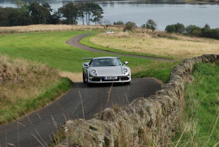 Sprints and Hillclimbs? - Page 1 - Noble - PistonHeads - In this dynamic image, a white sports car is the focal point as it races down a curvy road, leaning into a left turn. The road itself is quite scenic, flanked by a stone wall on its left side and an array of verdant trees on the right, providing a natural backdrop to the scene. In the distance, a serene lake mirrors the clear sky, reflecting hues of blue. A few other vehicles are present on the road, though they are dwarfed by the speeding sports car. This image captures the thrill of a sports car ride through a tranquil, natural landscape.