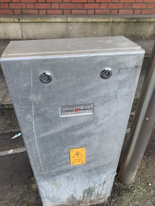 Life has been draining lately... - Page 159 - The Lounge - PistonHeads UK - The image shows a concrete utility box, situated on the sidewalk in front of a building with red brick walls. The utility box features two circular knobs on its side and has some stickers and writing on it. There is also a small yellow sign with a black arrow pointing upwards attached to the top right corner of the box. The overall scene suggests an urban setting, possibly indicating maintenance or utility services in the area.