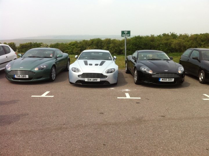 Pistonheads - The image displays a row of four distinct sports cars parked side-by-side. From left to right, the first car appears to be a green Bentley, the second a white Porsche, the third a sleek black convertible, and the fourth is of a similar black color, but the specific car model is not visible. The cars are parked in a parking lot with a blurred background that suggests an open, outdoor setting, possibly with a hill and greenery in the distance. A small sign, partially visible in the top right corner of the image, offers guidance for drivers. The overall scene suggests a scenic parking location, likely a popular stopping point for car enthusiasts.