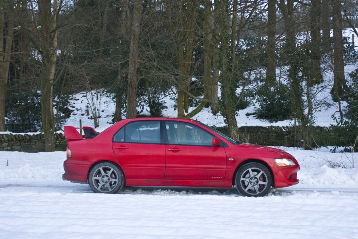 Pistonheads Show Motor - The image captures a scene of a vibrant red sports car parked amidst a picturesque, snow-covered landscape. The car, positioned in the middle left of the frame, is angled slightly to the left, showing its side and back profile. It's positioned between what appears to be a gate and a stand of trees, adding a sense of dynamism to the static scene. The ground around the car is blanketed in pristine white snow, while the trees adjacent to the car have their branches heavily laden with snowfall, suggesting that the photo was taken during or after a day of heavy snow. The image seems to evoke a sense of tranquility and serenity, as the car is the sole focal point in this expansive and natural setting.