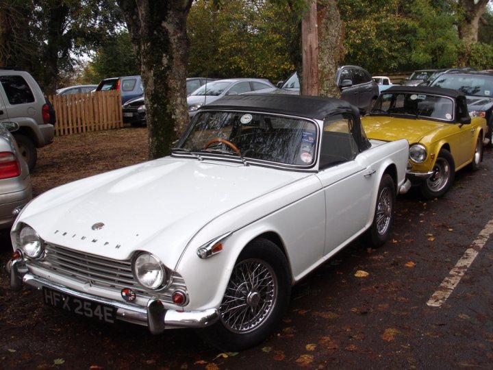 COOL CLASSIC CAR SPOTTERS POST!!! - Page 475 - Classic Cars and Yesterday's Heroes - PistonHeads - The image displays a vintage white car prominently parked among several other cars in a row. The vintage car has a classic design with round headlights, and it is adorned with stickers and a European license plate. There are trees in the background, suggesting this might be a parking lot area. The surrounding cars appear modern and are parked in a well-organized parking lot. The vintage car stands out due to its distinctive color and design.