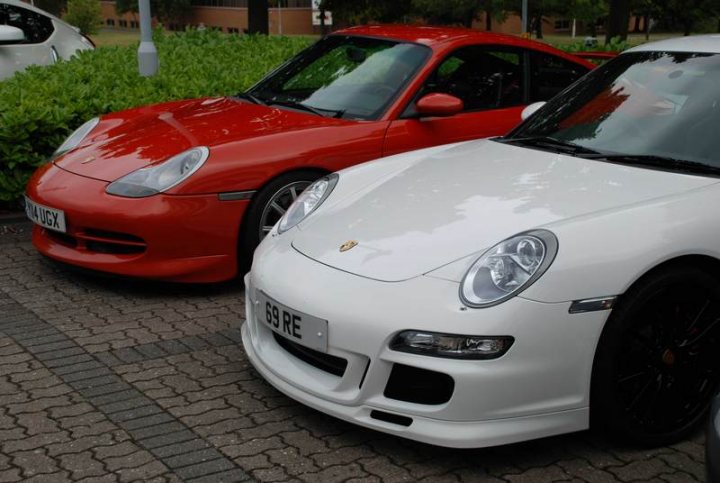 RE: PistonHeads Sunday Service: 25/07/10 - Page 9 - Events/Meetings/Travel - PistonHeads - This image showcases two sports cars parked side by side on a surface littered with pebbles. On the left is a vibrant red Porsche sports car, distinguished by a black stripe running along its length. Adjacent to it on the right is a sleek, white Volkswagen sports car, characterized by a smaller black stripe and silver accents. Both cars display a similar design language and appear to be of the high-performance variety. The background is a glimpse of greenery, suggesting that the location might be a parking area in a park or a similar outdoor setting.