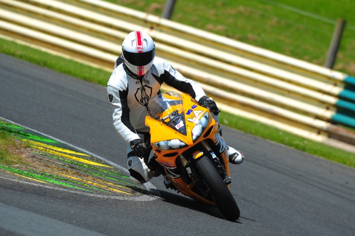 A man riding a motorcycle on a race track - Pistonheads - The image captures a dynamic scene from a motorcycle race. A rider, dressed in a full racing suit and riding a motorcycle painted in a vibrant yellow and orange color scheme, is in the midst of a turn on the race track. The rider appears focused and skilled, leaning into the curve with precision. The track itself is populated with barriers covered in green and yellow paint, indicating a professional racing environment. The surroundings include grassy areas and what appears to be a fence or other track infrastructure, all set against the backdrop of a clear day with blue skies.