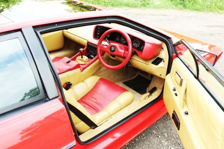 Pistonheads Pick Late - The image shows a red classic convertible car. The car's interior is visible, revealing a tan leather driver's seat adorned with a red steering wheel and numerous gauges. The passenger door is open, revealing a red seat and two black straps. The vehicle has no visible occupants, and it is parked on a surface that appears to be a dirt road or the side of a street. The hood of the car is up, exposing the engine area.