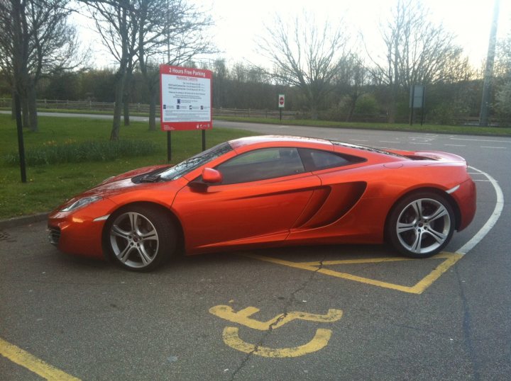 MP4-12C photos and review/comparison - Page 24 - Supercar General - PistonHeads - The image features a striking orange Lamborghini sports car parked in a parking lot. The car's sleek design and vibrant color make it the focal point of the image. Behind the car, there's a red sign with white text sitting on a grassy patch, which adds a pop of color to the scene. The car's license plate is also visible. A yellow handicap symbol is painted on the ground near the car, indicating that the lot is accessible for individuals with disabilities. The overall scene suggests a day of driving enjoyment.