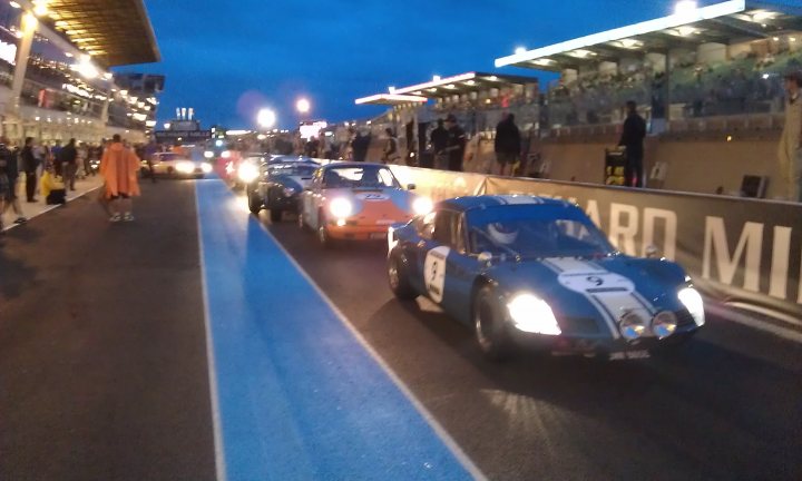 A group of people standing on the side of a road - Pistonheads - The image captures a vibrant automobile track at dusk. Numerous sports cars, each with a unique color and make, are in a procession driving on the track. The cars are illuminated with their headlights on, adding to the intensity of the scene. Spectators are present, scattered around the track, observing the show. The stands in the background, illuminated with a warm glow, provide an aesthetic backdrop to this lively scene.