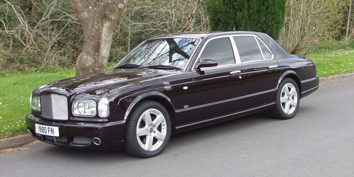 Buying a Bentley Arnage - Page 1 - Rolls Royce & Bentley - PistonHeads - The image displays a dark-colored, luxury sedan parked on a paved sidewalk alongside a tree with bare twigs in the background. The car features a large grille, sleek contours, and multi-spoke wheels that indicate its premium nature. The vehicle has a distinct front and side design that is characteristic of certain high-end brands, although the brand name is not visible. It appears to be in good condition, with no noticeable dents or scrapes. The setting seems to be in a residential or park-like area due to the presence of the tree and the natural, undisturbed background.