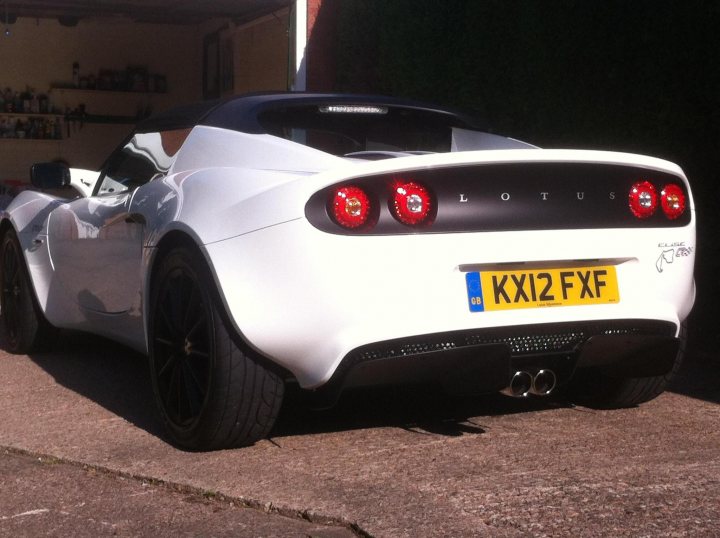 A car parked next to a parking meter - Pistonheads - The image features a parked white Lotus sports car, which is the centerpiece of the frame. The vehicle is adorned with a license plate reading "KXI2 FXF". The exhaust pipes, proudly displaying "7A", are prominently seen at the back of the car. The backdrop is a garage with a concrete floor, providing a solid contrast to the sleek lines of the car. The car, with its vibrant colors and sleek design, stands out vividly within this environment.