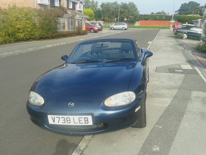 Got myself an MX-5 at last!! - Page 1 - Mazda MX5/Eunos/Miata - PistonHeads - The image depicts a single blue car parked on the side of a street. The car appears to be a modern convertible, characterized by its retractable hard top roof. Around it, the road is mostly clear, with a few other cars parked further down the street, suggesting this might be a suburban or residential area. On the side of the street, there's a well-maintained lawn and brick fence, contributing to the peaceful and clean neighborhood ambiance. The car itself is well kept and gleaming in the sunlight, adding to the overall pleasantness of the scene.