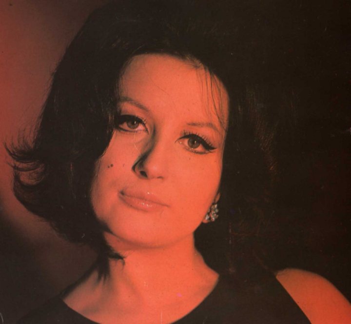 Mina - The image captures a portrait of a woman in what appears to be the 1960s or 1970s judging by her fashion and the overall color tone and style. She has a striking appearance with bold makeup, notably her eyes which are accentuated with dark eyeliner and thick eye shadows. The woman is wearing a geometric pattern on her clothing, which adds to her distinctive look. She has a classic short haircut complemented by stylish side-swept bangs. The lighting is dramatic, casting a deep red hue over the image, which brings a sense of sophistication to the overall portrayal.