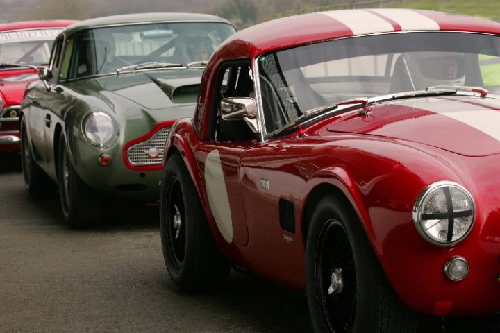 Masters race series, 10th March - Page 1 - Goodwood Events - PistonHeads - The image showcases a row of vintage cars, parked in a line along the side of a road. The cars are sleek and shiny, indicating that they are well-maintained. The lead car is a vibrant red convertible, and behind it, there are two green classic cars. The front of the fourth car in the line is visible, suggesting that the scene continues further back, possibly indicating a line of such cars. The presence of these cars on the street suggests that this could be a classic car show or gathering.