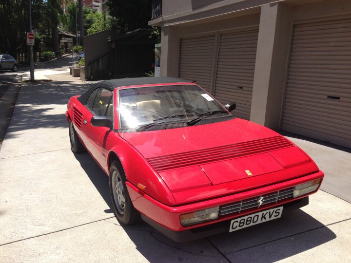 Wheel alignment specialist in Sydney - Page 1 - Australia - PistonHeads - In the image, there is a vibrant red Ferrari sports car parked next to a beige garage. The car is facing the viewer with its long, stylish hood prominently displayed. The license plate reads "C80 KVS". This high-end sports car is situated on a street lined with grass and trees, adding a touch of serenity to the scene. The backdrop of the car showcases a quaint house and another car parked further away. This scene paints a picture of luxury and elegance amidst a peaceful suburban setting.