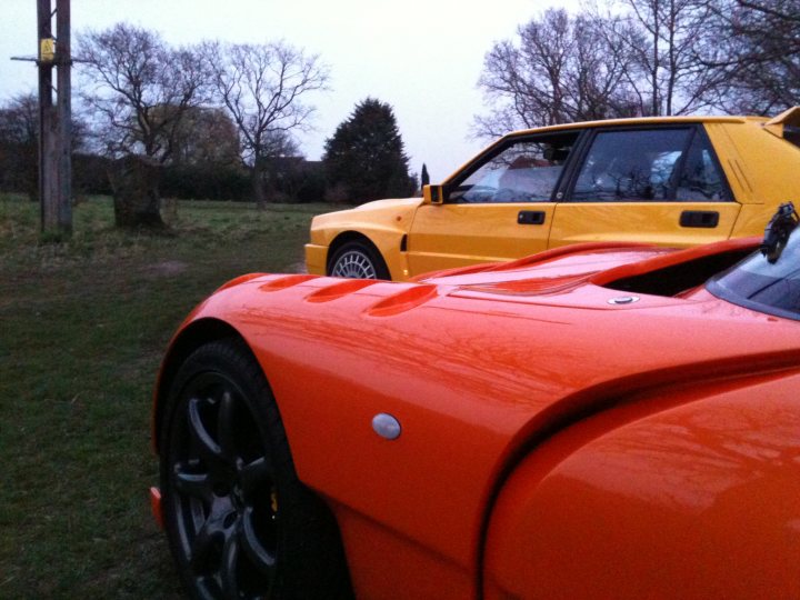 Pistonheads Seconds Endulge - The image shows an orange sports car parked on a grassy field near a yellow compact car with a vinyl top, suggesting a casual gathering or perhaps a competition given the presence of a track car. The sky is overcast, creating a muted, moody lighting that contrasts with the vivid hues of the vehicles. The grass has been worn in places, especially in the areas surrounding the cars, indicating frequent movement in that vicinity. Trees are visible in the background, adding a touch of nature to the scene. The image seems to capture a moment of quiet before or after some fast-paced driving.