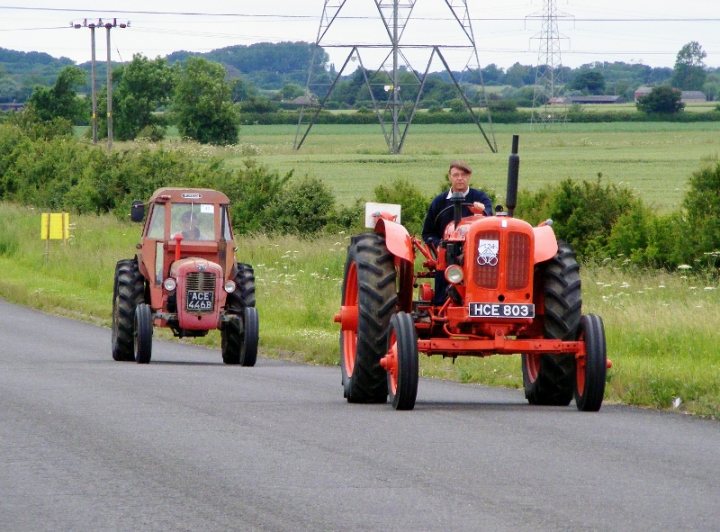 Herts, Beds, Bucks & Cambs Spotted - Page 332 - Herts, Beds, Bucks & Cambs - PistonHeads - In the image, there are two vintage tractors traveling down a road. The tractors have a distinct rustic charm, their bodies appearing well-maintained despite their age. The left tractor has red fenders, while the right one has orange ones, adding a splash of color to their otherwise monochrome appearance. The tractors are driving on both sides of the road, one on each side, indicating that the road is wide enough to accommodate their size. The background reveals a rural landscape with lush green grass and trees, hinting at a countryside location or possibly a tractor parade.