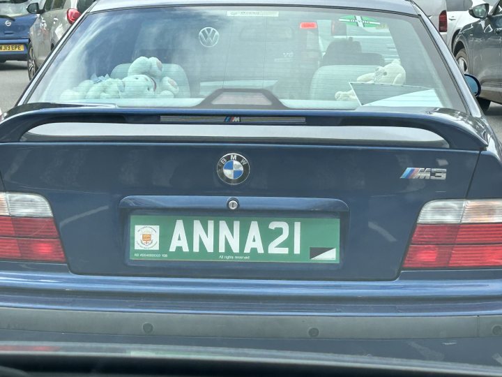 Identify this numberplate? - Page 1 - General Gassing - PistonHeads UK - The image features a car parked in what appears to be an outdoor parking area. On the back window of the car, there's a personalized license plate with the name "Anna 21" and another name that is partially obscured. In the back seat of the car, two teddy bears are visible. The car itself is dark in color, possibly black or a dark blue. There are no people visible in the immediate vicinity of the car. The surroundings suggest an urban setting with other cars parked nearby and possibly a street in view.