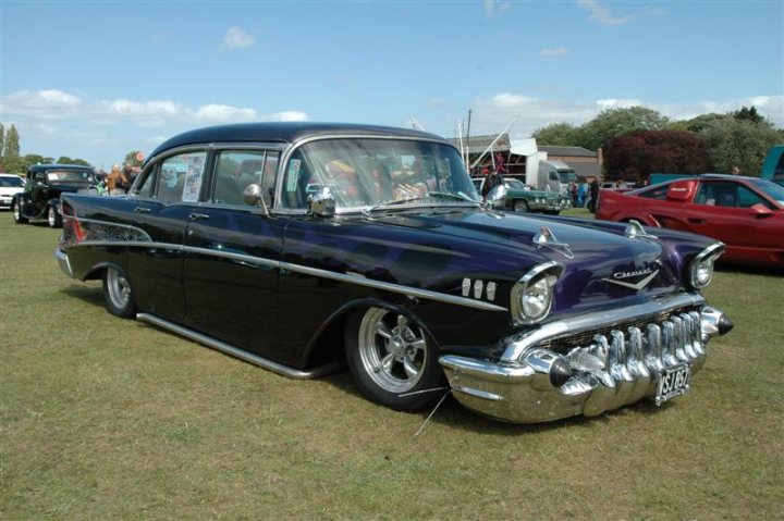 Fins and Chrome - Page 1 - North East - PistonHeads - The image showcases an antique car display at an outdoor setting, likely a classic car show. The car that's the main focus is a dark blue Chevrolet Nomad, a vintage model from the 1950s, characterized by its two fins. It's parked on grass, facing the right side of the image, with other cars and people in the background, under a clear blue sky with some clouds.